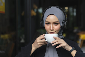 Close up portrait of Muslim woman in hijab enjoying coffee while wearing headphones. Relaxed modern...