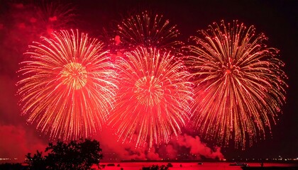 Bright, vibrant display of three red fireworks bursting in the night sky, with a silhouette of trees and water in the foreground