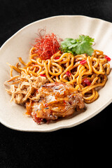 Stir-fried udon noodles with chicken, vegetables, and garnished with cilantro and chili threads on a beige plate. High angle view against a dark background.