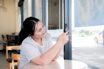 Asian woman smiling while taking photo with smartphone in a coffee shop near window. Relaxed lifestyle moment showing technology use, social media connection, and leisure time enjoyment.