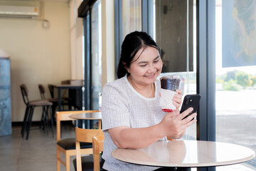 Asian woman smiling while using smartphone and holding iced drink in a coffee shop. Relaxed mood showing lifestyle, social connection, and modern communication during leisure time.