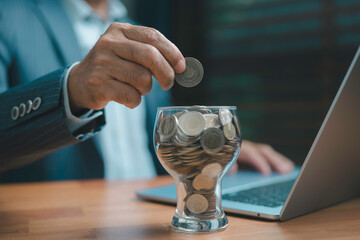 Businessman saving money by placing coins into a glass jar beside a laptop, symbolizing financial...