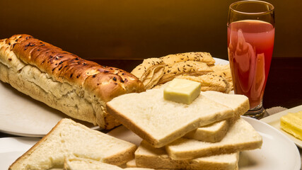 A warm and inviting close-up image of a breakfast table featuring freshly baked golden-brown bread loaf, white bread slices topped with a cube of butter, puff pastries and a glass of fresh fruit juice