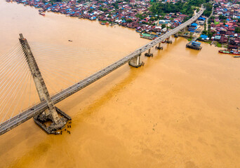 An aerial view of the Mahkota II Bridge crossing the Mahakam River in Samarinda, Borneo. The modern cable-stayed bridge spans over a dense residential settlement on the riverbank