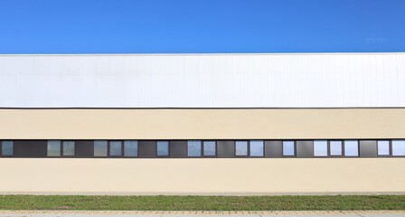 Modern yellow and  white industrial building with a rows of windows. Footpath, grass and street in front. Background for copy space.