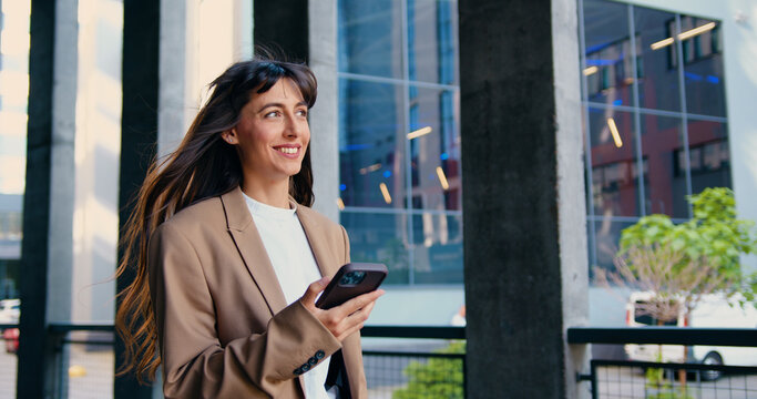 Confident and smiling young businesswoman walking outside modern office building, smiling while using smartphone, checking messages, browsing apps, staying connected on the go, outdoor