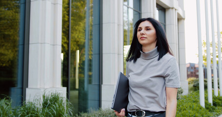 Portrait of successful brunette woman with laptop in hands, walking in modern business district.