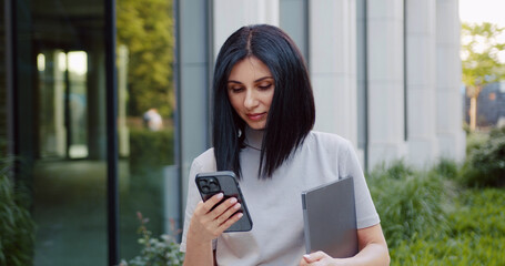Young caucasian business woman using cell phone mobile app and holding computer device for work, trading, chat with ai, working online. Outdoors