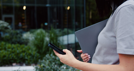 Side view of close up females hands using smartphone and holding laptop. Fingers hands dialing number on smartphone screen. Outdoors