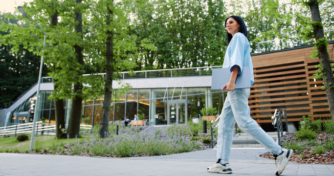 Confident young woman in casual clothes is walking through a city park holding laptop, returning to work or school, with green trees and modern glass buildings in the background on sunny day