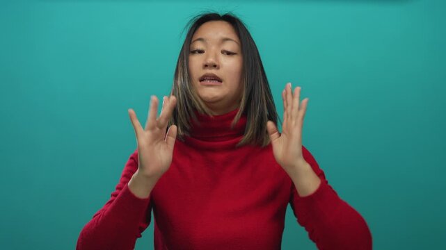 Woman gesturing for calm against an isolated green wall, wearing red sweater, with a serene expression and a youthful chinese appearance.