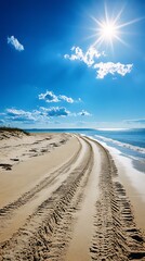 Sunlit sandy beach with tire tracks leading towards the ocean under a bright blue sky