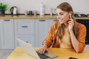 Young happy housewife IT woman in orange shirt casual clothes hold use work on laptop pc computer listen music in earphones sit at table in light kitchen at home alone Lifestyle cooking food concept