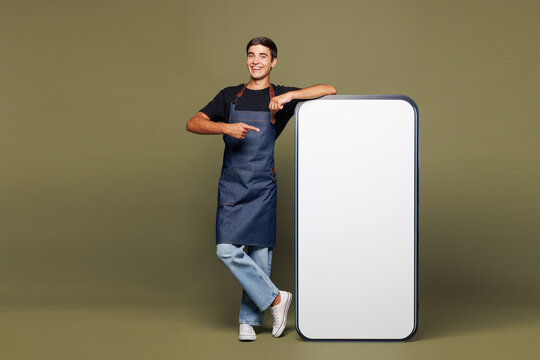Full body young man barista bartender barman wear apron t-shirt work in coffee shop point on big huge blank screen mobile cell phone isolated on plain green background. Small business startup concept.