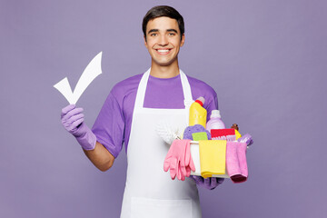 Young happy man wear violet t-shirt hold basin with detergent bottles while doing housework tidy up show check mark to do list isolated on plain pastel light purple background. Housekeeping concept.