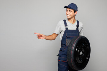 Young car mechanic man wear overall hat uniform workwear clothes work in garage hold wheel change tire point index finger aside on area isolated on plain grey background Automotive repair job concept