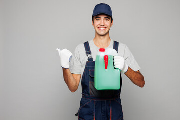Young car mechanic man wear overall hat uniform workwear clothes work in garage holding in hand bottle of coolant point finger aside isolated on plain grey background. Automotive repair job concept.