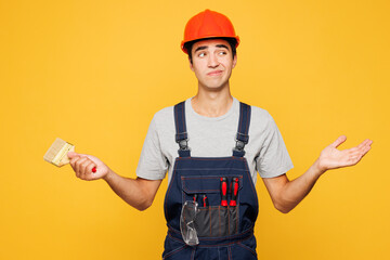 Young confused employee laborer handyman man wear helmet overall hold in hand paint brush isolated on plain yellow background. Instrument accessories for renovation apartment room Repair home concept