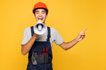 Young employee laborer man wear helmet overall scream in megaphone announces discounts sale point...