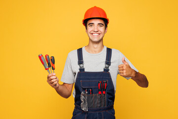 Young employee laborer handyman man wear helmet overall hold screwdrivers show thumb up isolated on plain yellow background. Instruments accessories for renovation apartment room. Repair home concept.