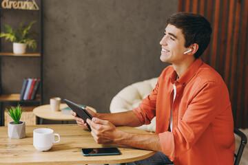 Young IT man he wear orange casual clothes digital tablet pc computer sitting at table in coffee shop cafe relax rest in restaurant during free time indoors. Freelance mobile office business concept.