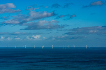 line of offshore wind turbines standing in calm blue sea beneath a bright sky with scattered clouds