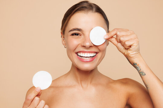 Beautiful happy half naked topless young woman 20s with nude make up hold cover eye with cotton pad isolated on plain beige background studio portrait Skin care healthcare cosmetic procedures concept
