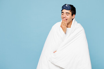 Calm young man wear pyjamas jam sleep eye mask rest relax at home wrapped in duvet blanket cover mouth with hand yawn isolated on plain pastel light blue cyan background. Good mood night nap concept.