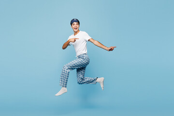 Obraz premium Full body calm young man wear pyjamas jam sleep eye mask rest relax at home jump high run fast point index finger aside isolated on plain pastel light blue cyan background Good mood night nap concept