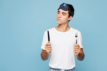 Full body calm young man he wear pyjamas jam sleep eye mask rest relax at home hold in hand knife and fork going to eat isolated on plain pastel light blue cyan background Good mood night nap concept