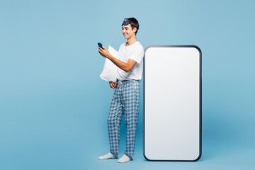 Full body calm young man he wear pyjamas jam sleep eye mask rest relax at home hold pillow big huge blank screen use mobile cell phone isolated on plain blue background. Good mood night nap concept.