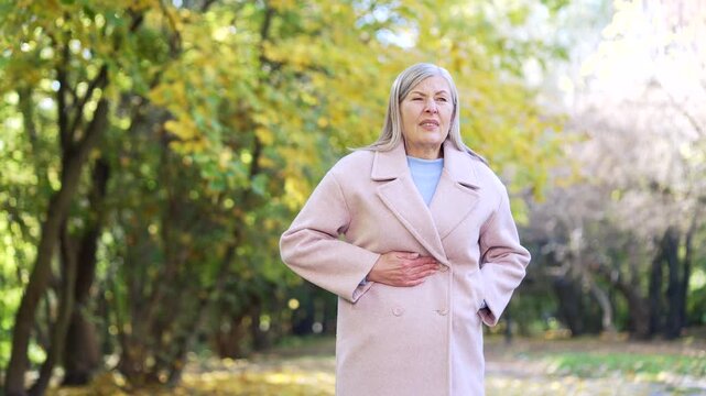 Senior woman suffering from stomach pain cramps standing outside in park. Mature eldery female outdoors Feels ache of internal organs, indigestion, poisoning, menstrual abdomen, pancreatitis gastritis