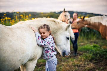Equine assisted therapy with child and horses on animal farm. Little girl embracing white pony...