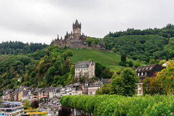 Cochem, Germany - September 9, 2025: Panoramic view of historic Reichsburg Cochem castle overlooking the Moselle River and old town, with river cruise boats on a cloudy autumn day.