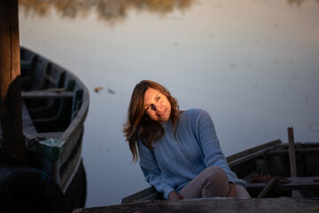 Portrait of young woman in blue sweater sits on an old wooden boat at sunset, smiling softly. Warm golden light highlights her face and hair, creating a calm, romantic atmosphere by the water.