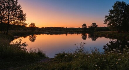 Fototapeta premium Still lake reflects warm sunset colors, framed by trees and wildflowers