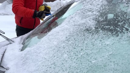 Man wearing orange winter coat scraping snow and ice from car windscreen on a snowy morning - Powered by Adobe