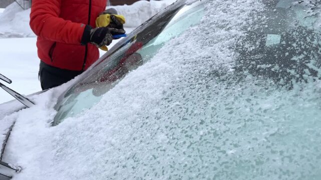 Adult man wearing orange coat scraping ice from car windscreen on winter morning