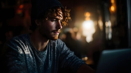 Young man with curly hair intently working on a laptop in a dimly lit atmospheric cafe setting with bokeh lights