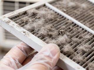 A hand in a protective glove holds a severely clogged air filter thick with gray dust and debris, highlighting maintenance.