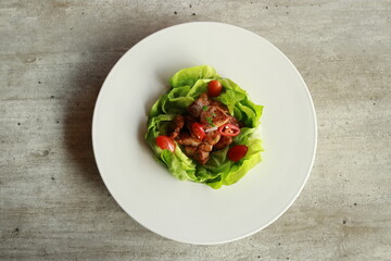 A culinary delight featuring a dish of succulent meat nestled within fresh lettuce leaves and complemented by ripe cherry tomatoes, all elegantly presented on a pristine plate.