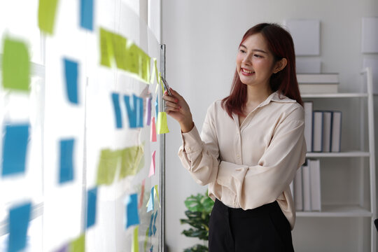 Young businesswoman is using colorful sticky notes on a glass board, organizing her thoughts and ideas for a successful business strategy in office.