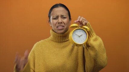 Hispanic woman in a yellow sweater holds a yellow alarm clock against an orange background, expressing frustration over time management issues.