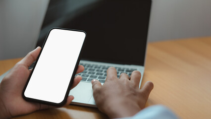 Hand Holding Smartphone with Blank Screen and Laptop on Desk for Mockup or Digital Business Concept