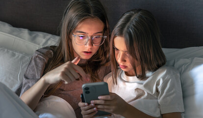 Two girls discuss something on a phone in bed. Shared scroll.