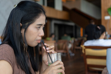 Asian woman holding a glass of green drink after sipping, straw no longer in her mouth, sitting at a restaurant table with bright natural light.