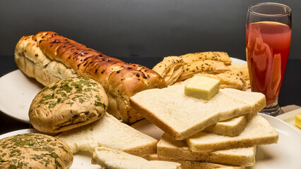 A close-up photo of a wholesome breakfast setup featuring freshly baked golden-brown bread loaf, white bread slices topped with butter, crisp puff pastries, and a glass of fruit juice.
