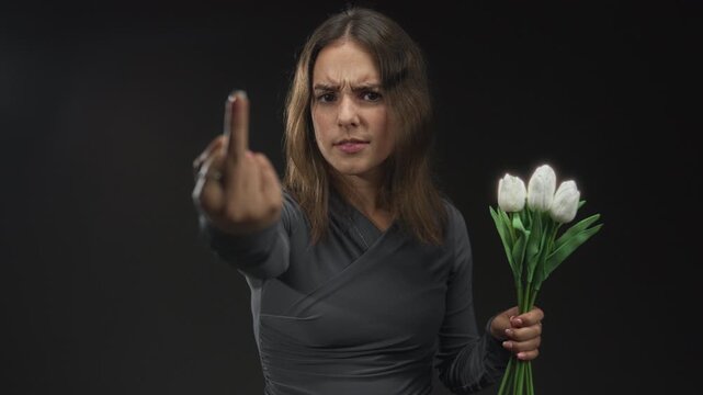 Young hispanic woman showing middle finger while holding white tulips in black studio; anger defiance.