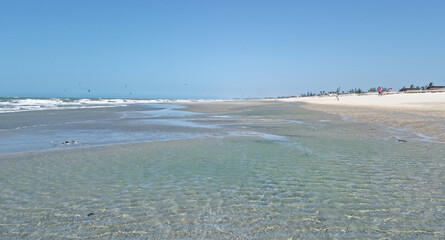 Calm tidal pools stretch along a sunlit beach, with soft waves, white sand, and distant kites...