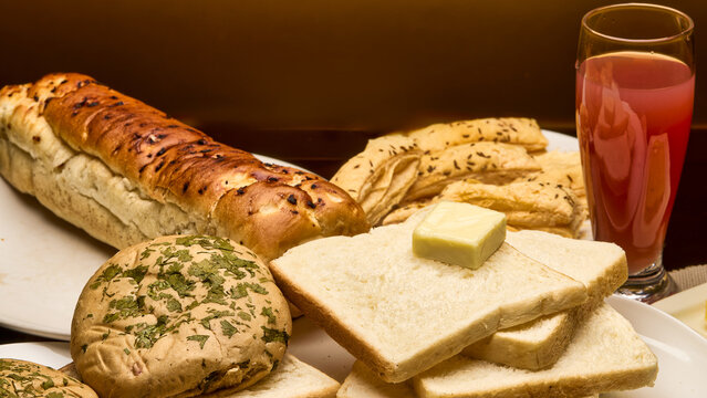 A beautifully composed image of assorted bakery items including buttered bread slices, buns, puff sticks, and a glass of fruit juice, set against a warm golden background